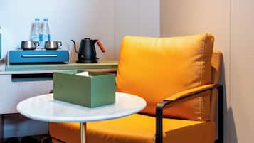A compact room with a single yellow chair, a small round table with a tissue box, a blue suitcase, a kettle, and two water bottles on a counter.