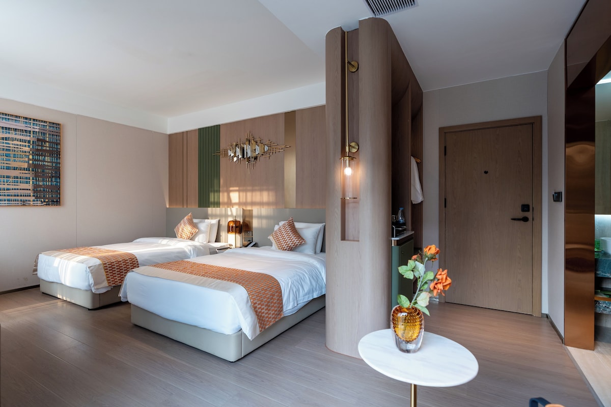 Una habitación de hotel moderna con dos camas, un espejo grande, una lámpara fijada a la pared y una mesita de noche con un florero.
