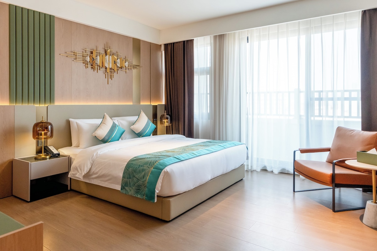 Habitación de hotel con una cama grande, mesitas de noche, una silla y vistas al exterior a través de cortinas translúcidas.