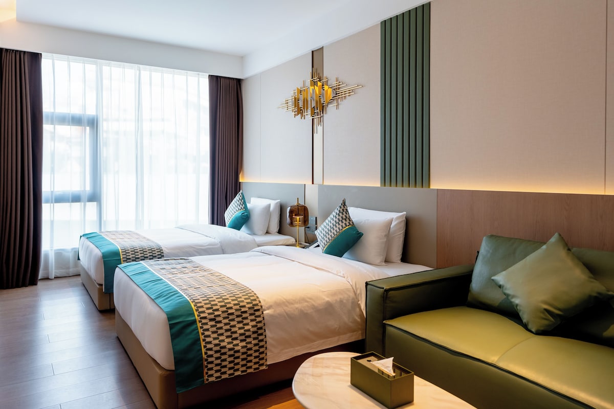 Habitación de hotel con dos camas, un sofá, una mesita de noche y un ventanal con cortinas.