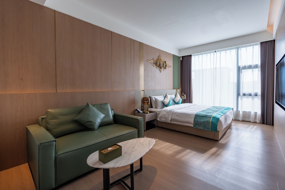 Una habitación de hotel moderna con un sofá verde, una cama con colcha blanca y aguamarina, una mesita con una caja y un ventanal con cortinas.