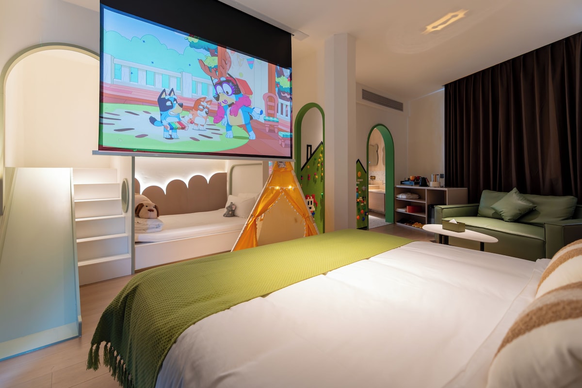 Una habitación de hotel con una cama, televisor, sofá verde y un área de juegos con una tienda de campaña.