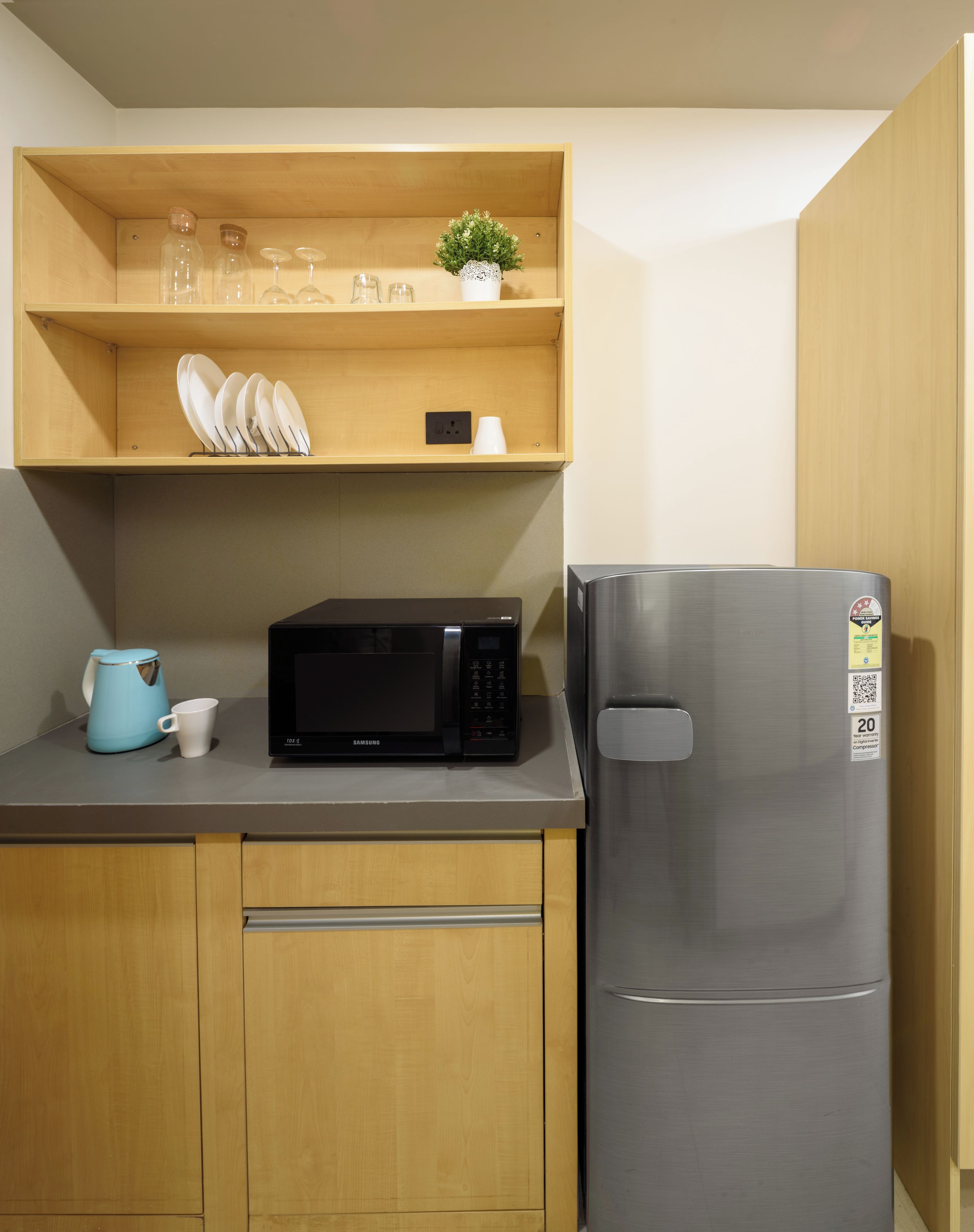 A compact kitchen with a microwave, a stainless steel refrigerator, wooden cabinets, and a shelf with glassware and a small plant.