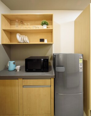A compact kitchen with a microwave, a stainless steel refrigerator, wooden cabinets, and a shelf with glassware and a small plant.