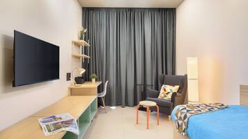 Una habitación de hotel moderna con televisor de pantalla plana, un escritorio con lámpara, una silla, una cama y un ventanal con cortinas.