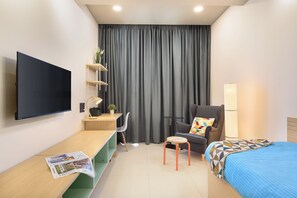 Une chambre d’hôtel moderne équipée d’une télévision à écran plat, d’un bureau avec une lampe, d’une chaise, d’un lit et d’une grande fenêtre avec des rideaux.