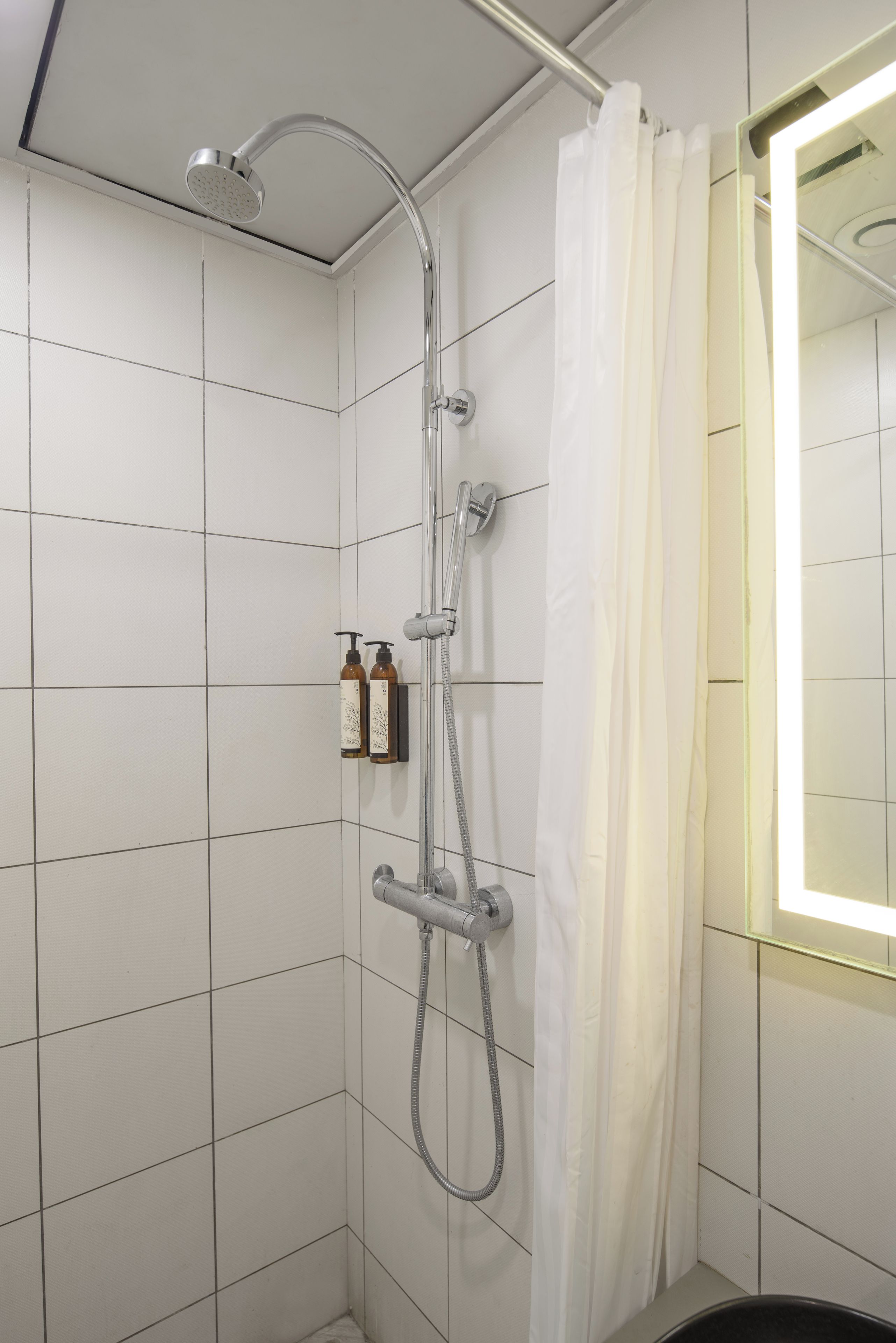 A shower with a handheld showerhead and a wall-mounted faucet, a curtain, and a mirror reflecting the shower area.