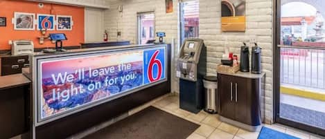 A motel lobby with a reception desk, a digital sign, a coffee machine, and a view of the parking area.