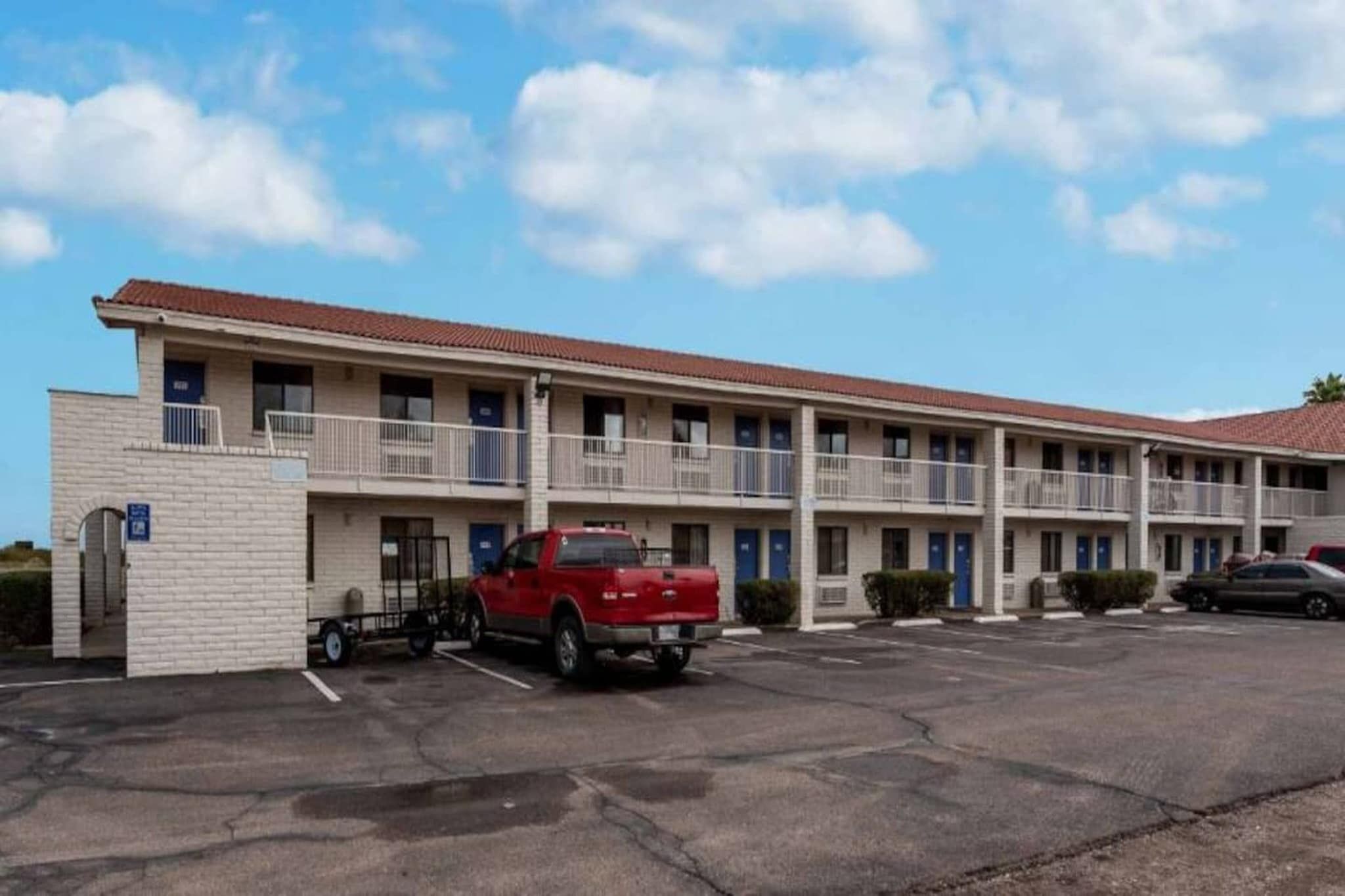 A multi-story motel with a parking lot, a red pickup truck, and a clear blue sky.