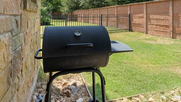 Una parrilla de barbacoa ubicada frente a una pared de piedra, con un césped y una cerca de metal al fondo.