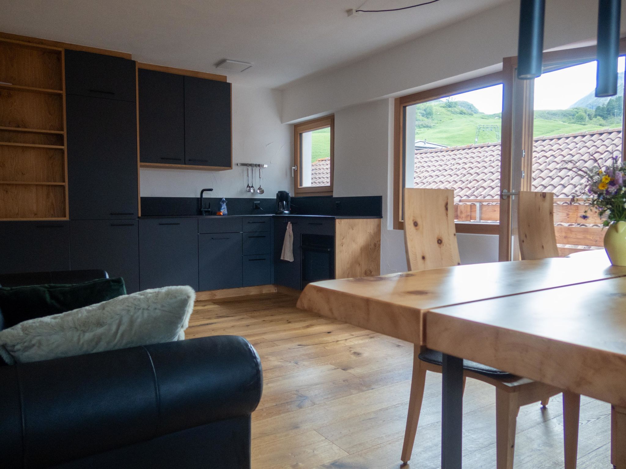 A modern kitchen with dark cabinets, a wooden dining table, and a view of a green landscape through large windows.