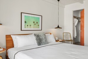 Un lit bien fait, avec du linge de lit blanc, un tableau encadré représentant des palmiers, une lampe suspendue et une chaise.
