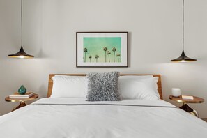 A neatly made bed with a white pillow, a framed picture of palm trees, and two pendant lights. - The Weekend by AvantStay | 3 Suites • Boutique Stay for 12, Private Patios, Pool (Palm Springs)