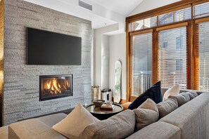 A modern living room with a fireplace, a flat-screen TV mounted on a textured wall, and a large window with blinds. - Mountain Views | Pool & Spa | Walk to Gondola | Village Dining | The Iron Horse (Truckee)