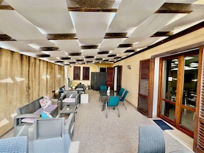 A covered outdoor seating area with tables and chairs, a wooden wall, and a large sliding glass door. - Vrbo Property (Marsala)