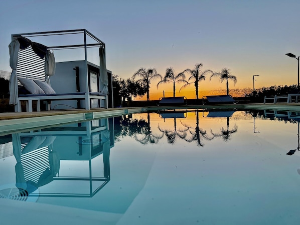 A clear pool with lounge chairs and a shaded area, palm trees, and a sunset in the background. - Vrbo Property (Marsala)