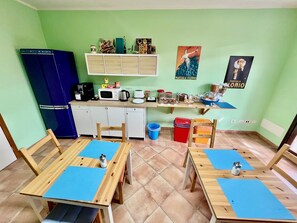 Een kleine kitchenette met een magnetron, koffiezetapparaat en kastjes. Er staan twee houten tafels met blauwe tafelonderleggers en stoelen.
