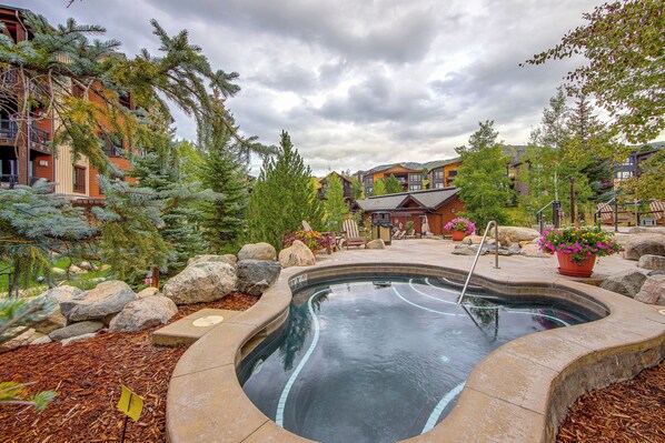 A swimming pool with a stone border, surrounded by rocks and potted plants, with buildings in the background. - Gondola to Skiing! Steamboat Condo w/ Hot Tubs (Steamboat Springs)