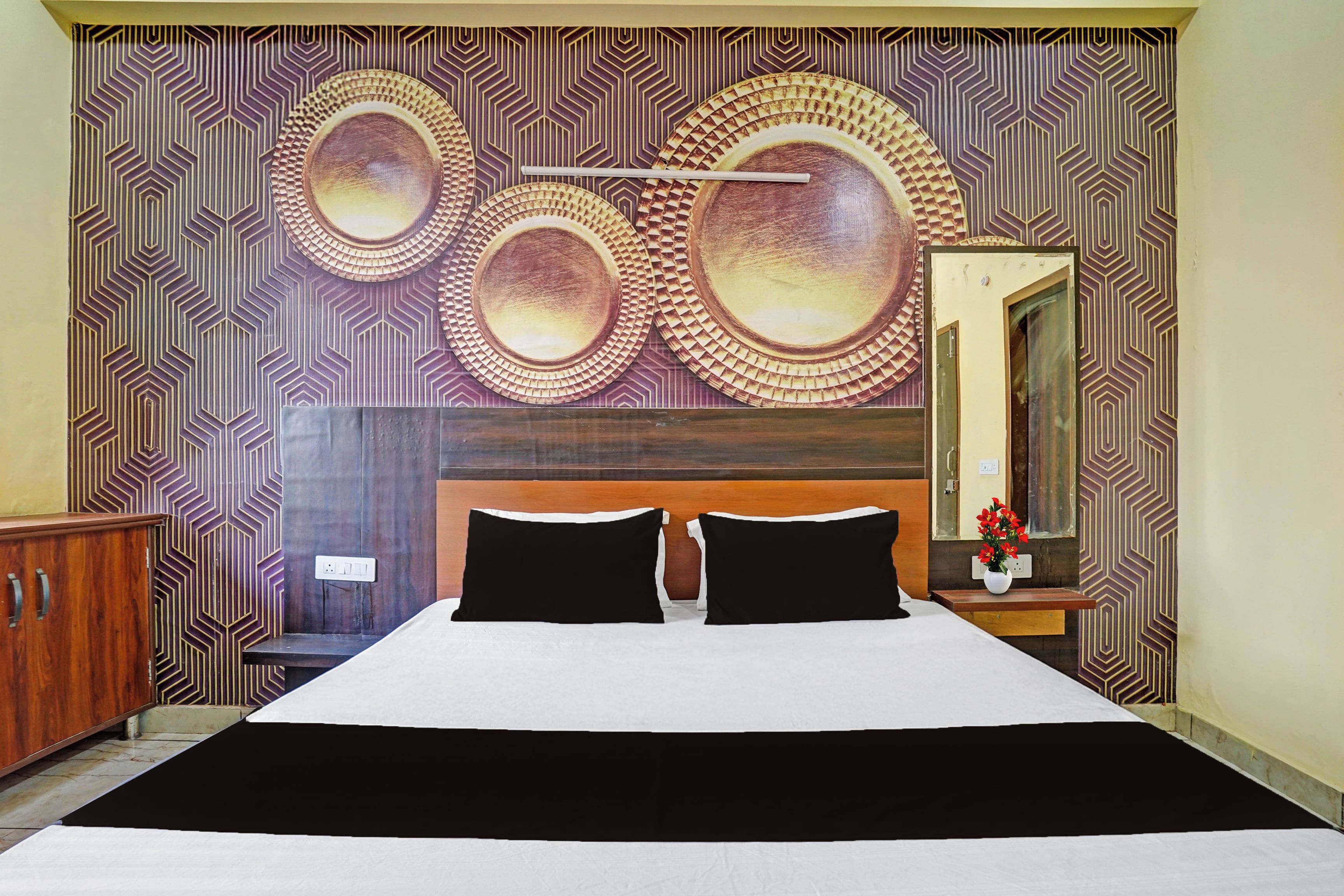 A hotel room with a large bed, two black pillows, a wooden headboard, a wall with circular decorative patterns, and a mirror with a vase of flowers.