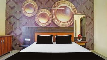 A hotel room with a large bed, two black pillows, a wooden headboard, a wall with circular decorative patterns, and a mirror with a vase of flowers.