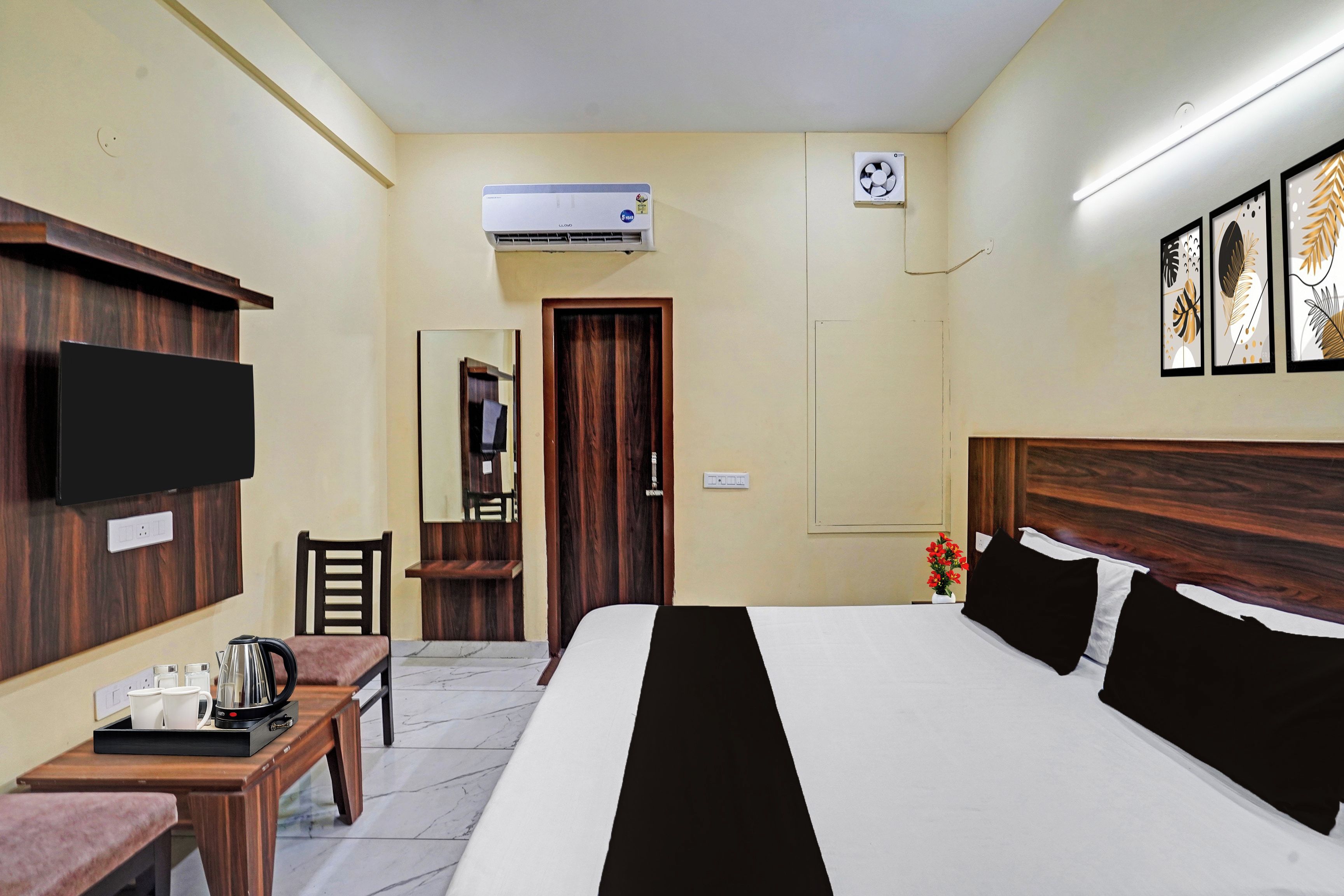 A hotel room with a bed, a desk with a kettle, a chair, a TV, an air conditioner, and framed artwork on the wall.