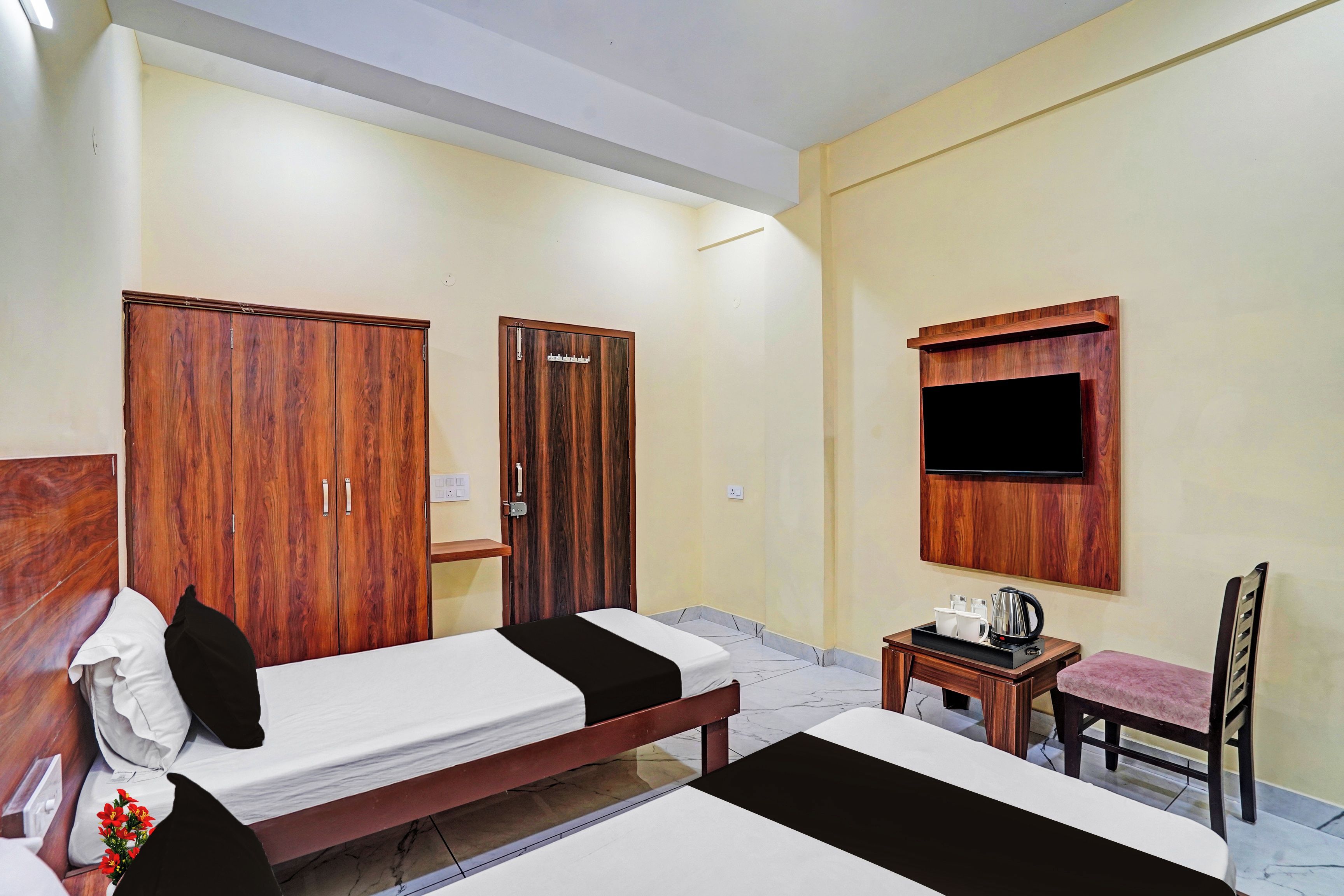 A hotel room with two beds, a wooden wardrobe, a small table with a kettle, and a television mounted on the wall.