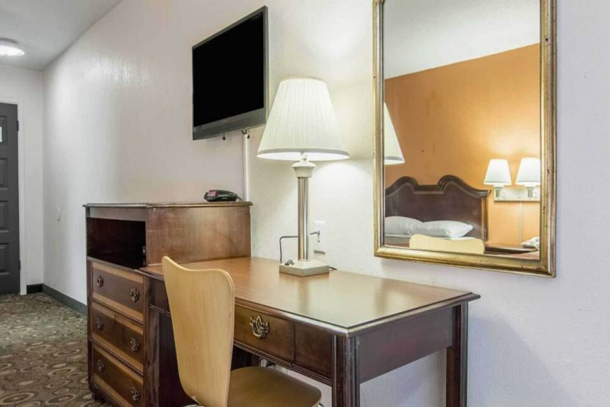 Habitación de hotel con un escritorio de madera, una silla, un televisor de pantalla plana montado en la pared, una lámpara de noche y un espejo grande que refleja la habitación.