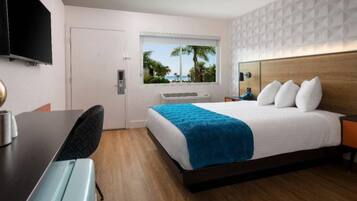 A hotel room with a bed, a desk, a chair, a TV, and a window with a view of palm trees and the sea.