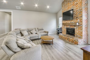 A living room with a sectional sofa, a wooden coffee table, and a brick fireplace with a flat-screen TV. - Play & Unwind! Home w/ Fenced Yard in Amarillo (Amarillo)