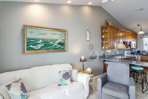 A living room with a sofa, a chair, a painting of the ocean, wooden cabinets, and a kitchen area with a sink and stove. - Unwind Poolside, Walk to Beach! Pensacola Getaway (Pensacola)