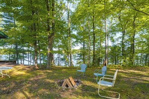 En campingplats vid sjön med två metallstolar, en eldstad och en träbänk.
