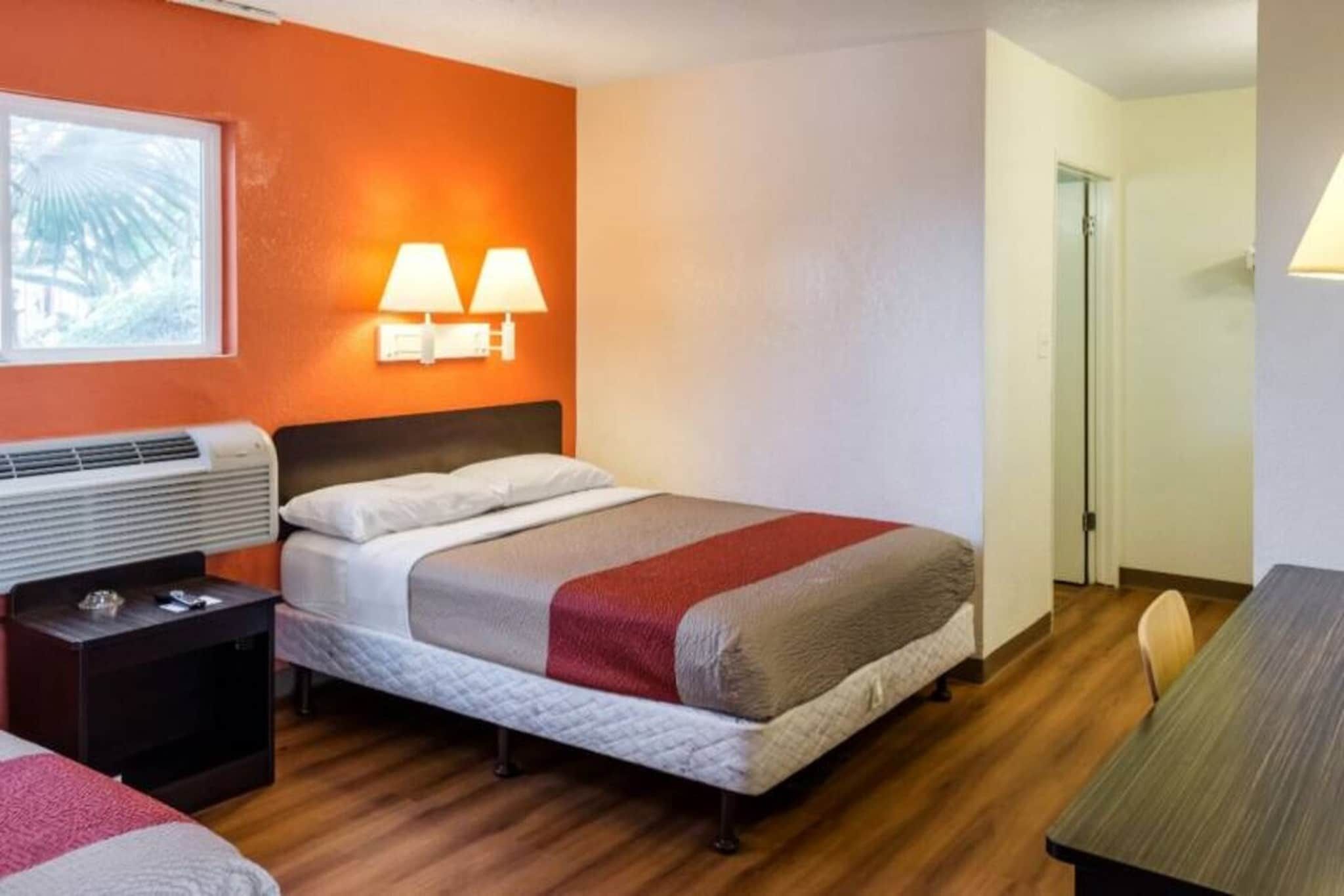 A hotel room with a bed, bedside table, lamp, and a view of the outside.