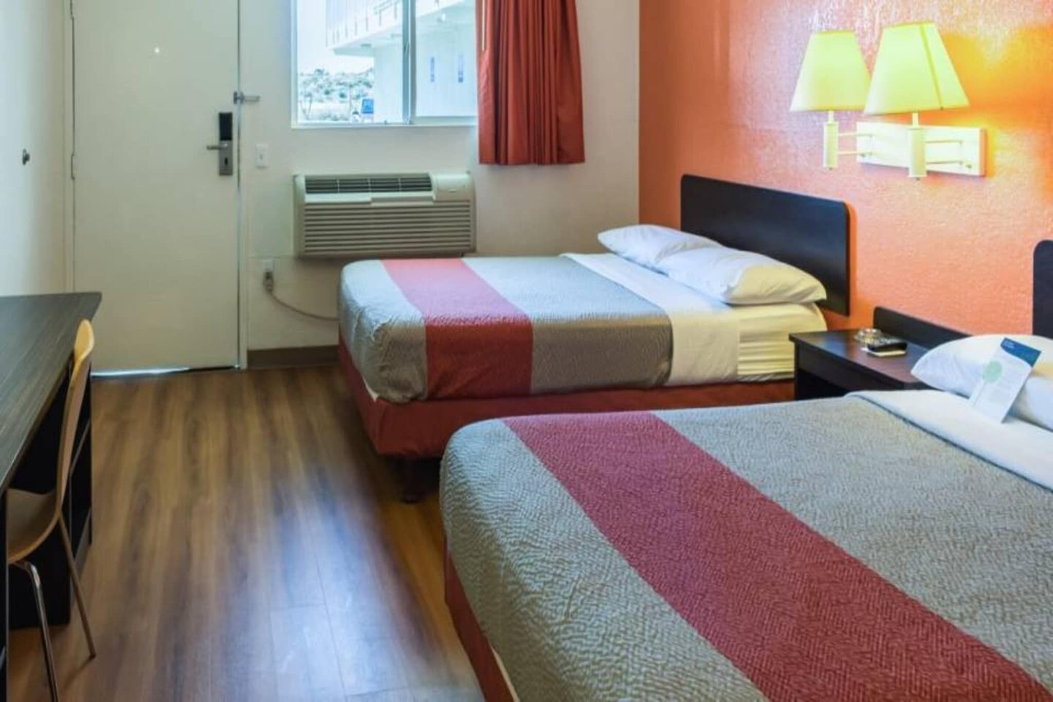 A hotel room with two beds, a desk, a chair, a window with curtains, an air conditioning unit, and wall-mounted lamps.