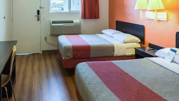 A hotel room with two beds, a desk, a chair, a window with curtains, an air conditioning unit, and wall-mounted lamps.