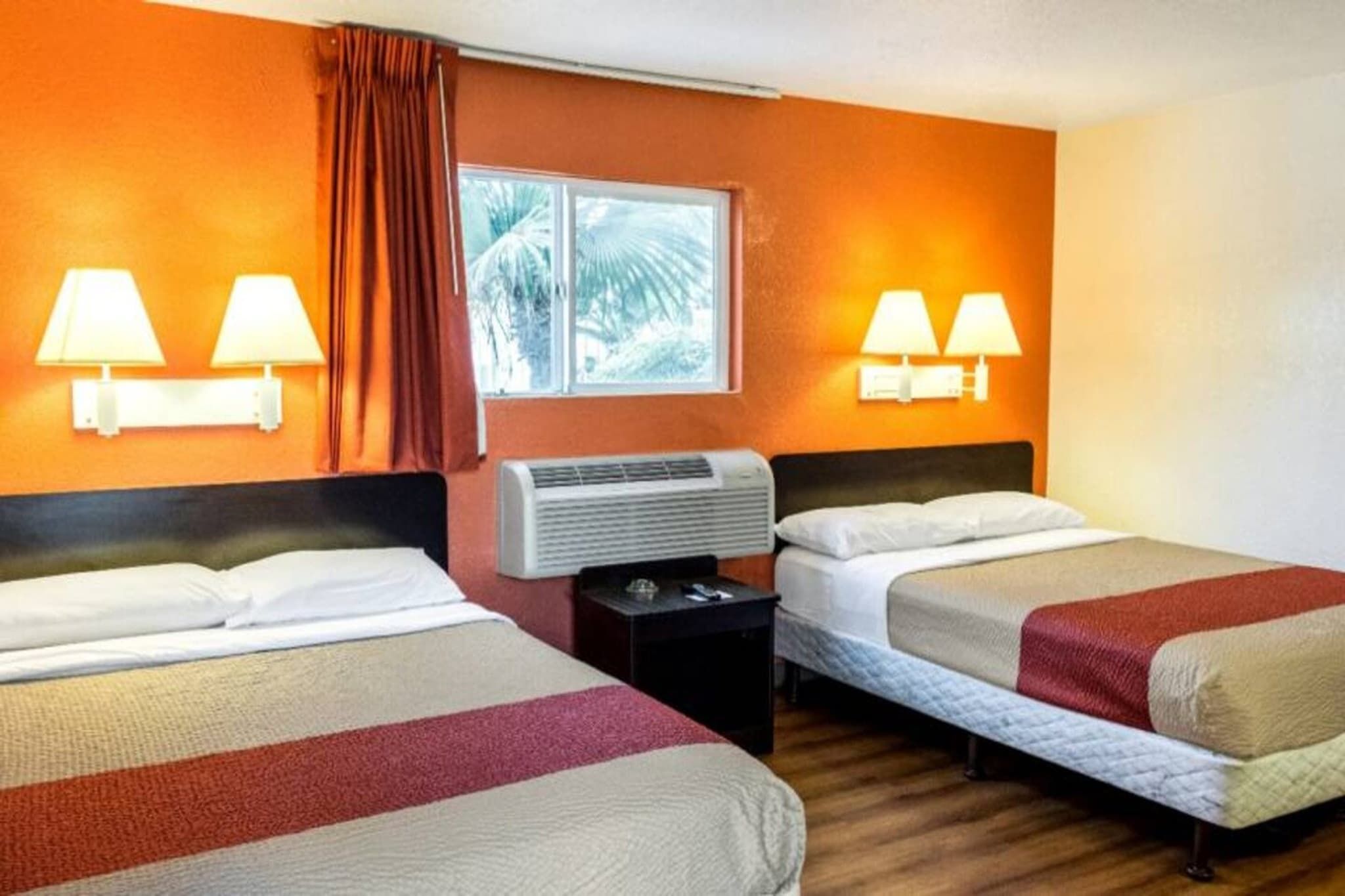 A hotel room with two beds, a window with curtains, wall-mounted lamps, and an air conditioning unit.