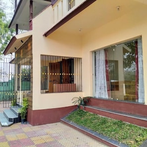 A two-story building with a balcony, a window with a curtain, and a small garden area.