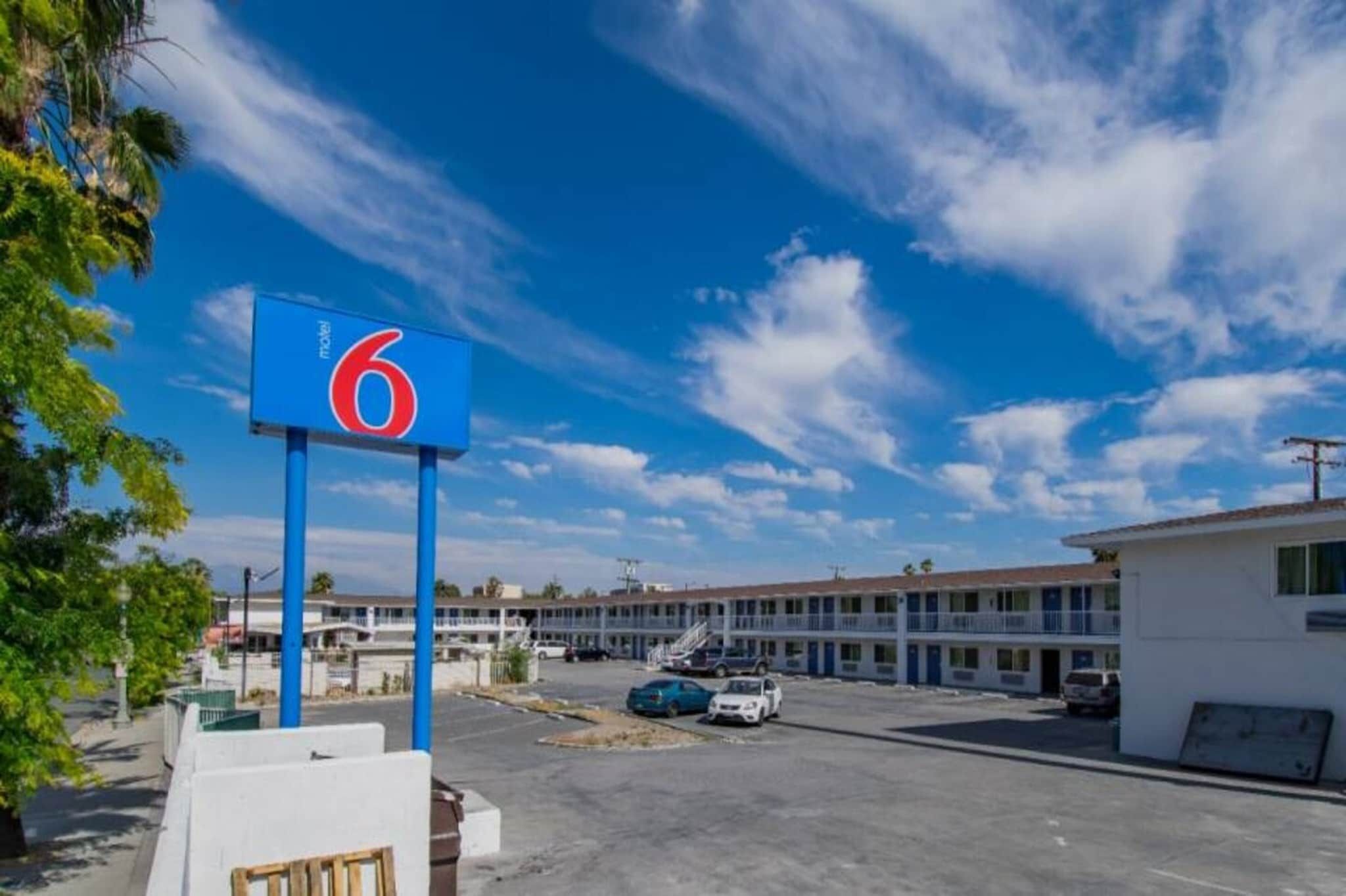 Un motel avec une enseigne bleue bien visible, sur laquelle figure le chiffre 6, un parking avec plusieurs voitures et une rangée de bâtiments du motel en arrière-plan.