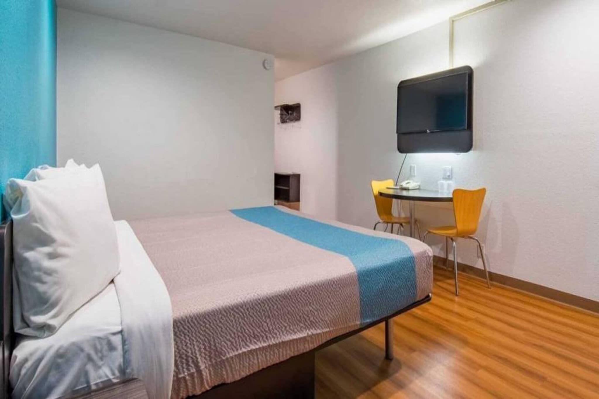 A hotel room with a bed, a desk, two chairs, a flat-screen TV, and a small shelf.