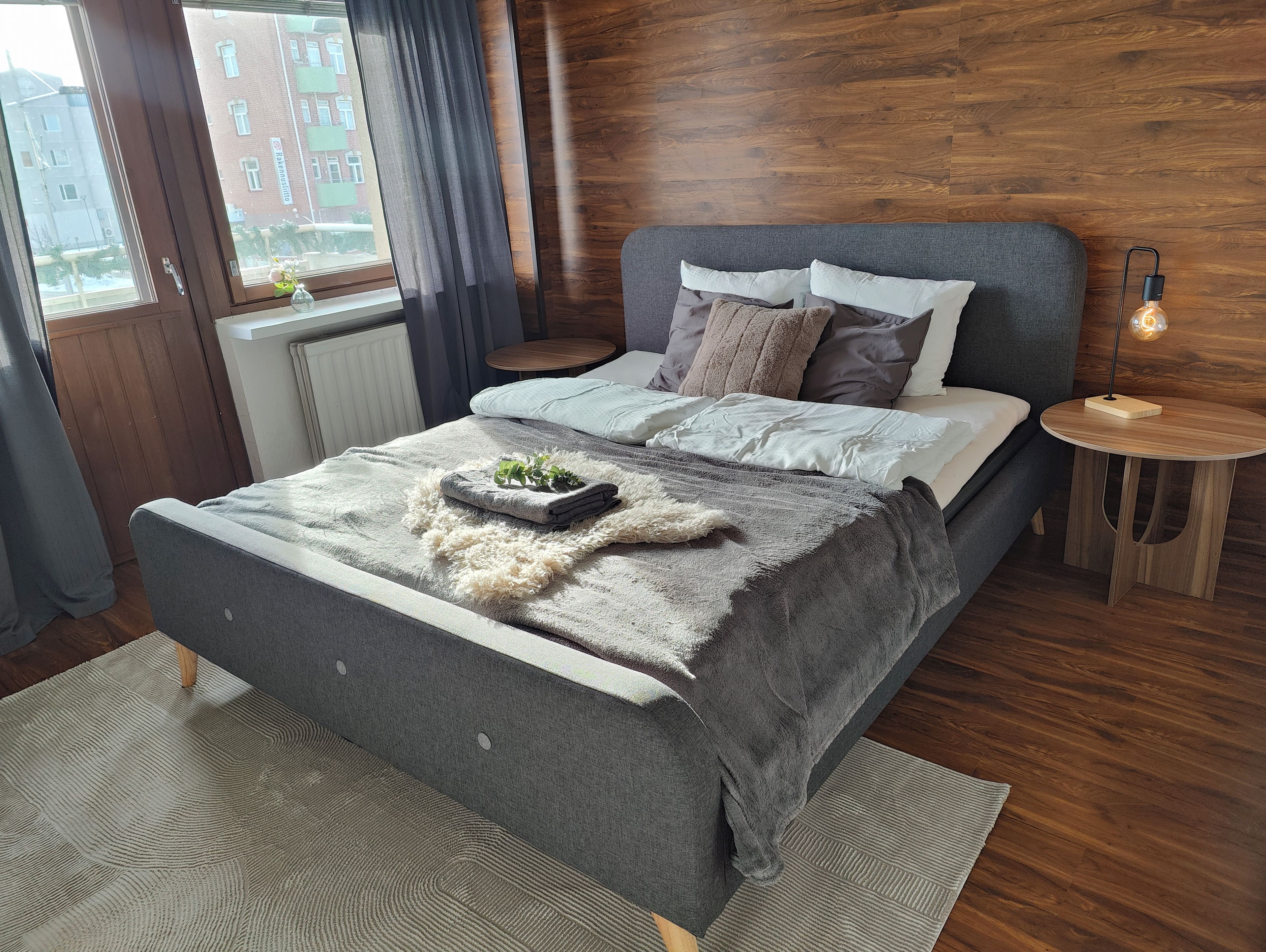 Une chambre à coucher moderne avec un lit gris, une tête de lit en bois et une petite table de chevet avec une lampe.