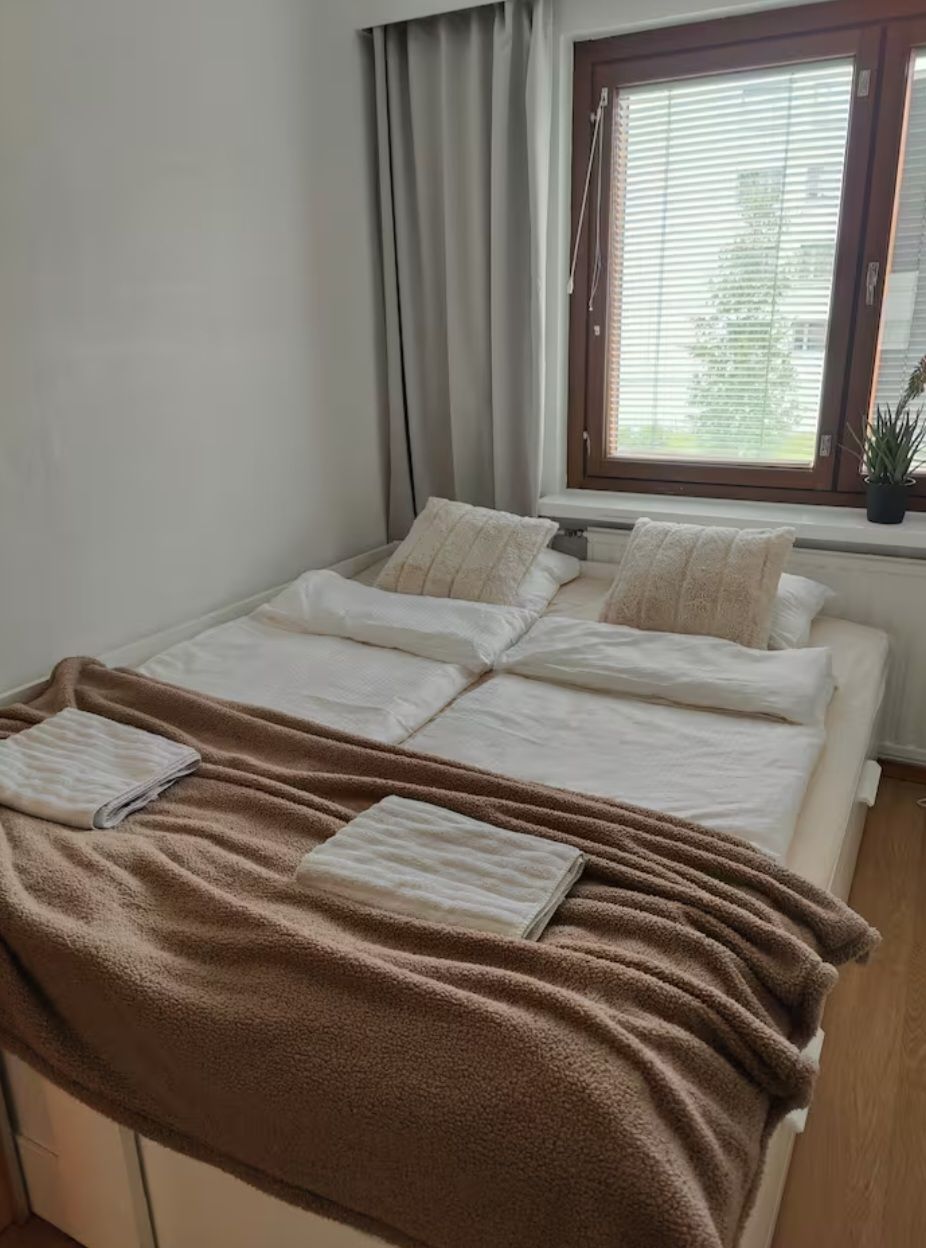 A neatly made bed with a brown blanket and pillows, a window with blinds, and a potted plant on the windowsill.