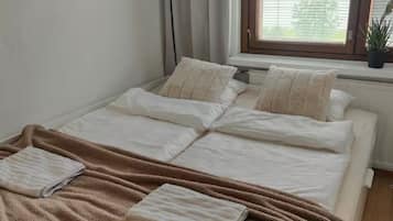 A neatly made bed with a brown blanket and pillows, a window with blinds, and a potted plant on the windowsill.