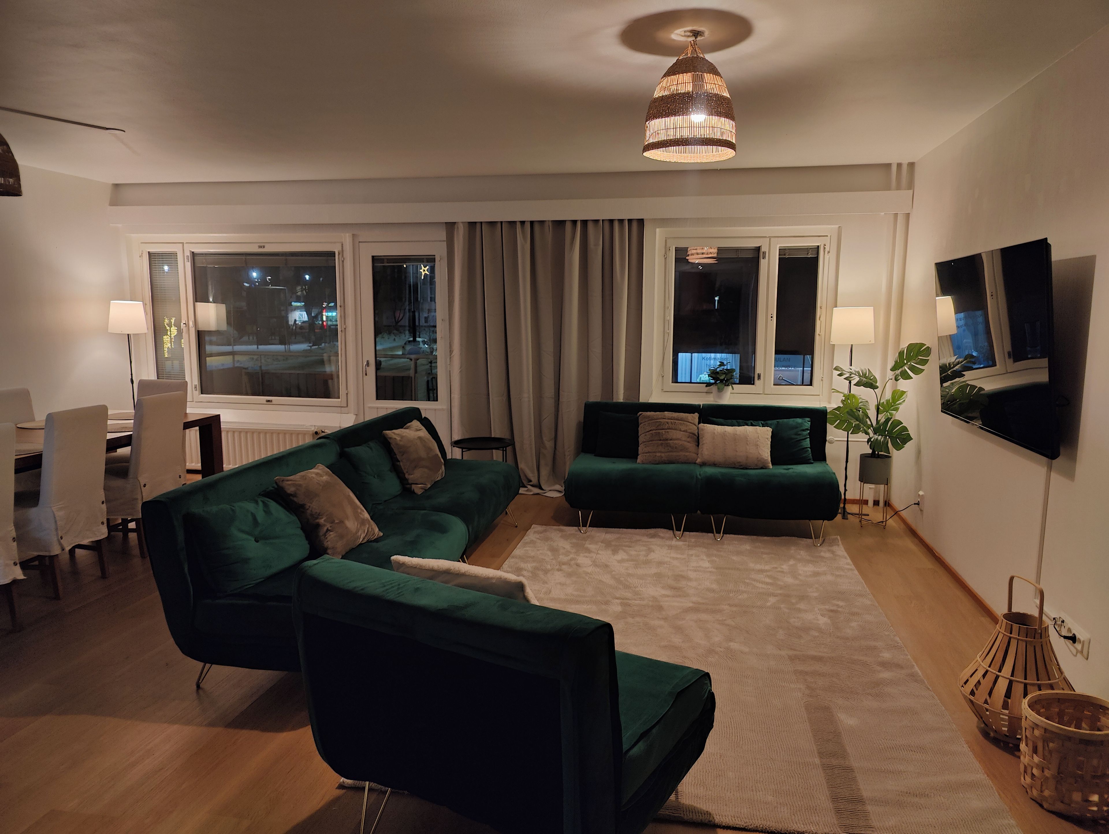 Ein modernes Wohnzimmer mit einem grünen Ecksofa, einem Esstisch mit Stühlen, einem an der Wand montierten Fernseher und einem großen Fenster mit Vorhängen.