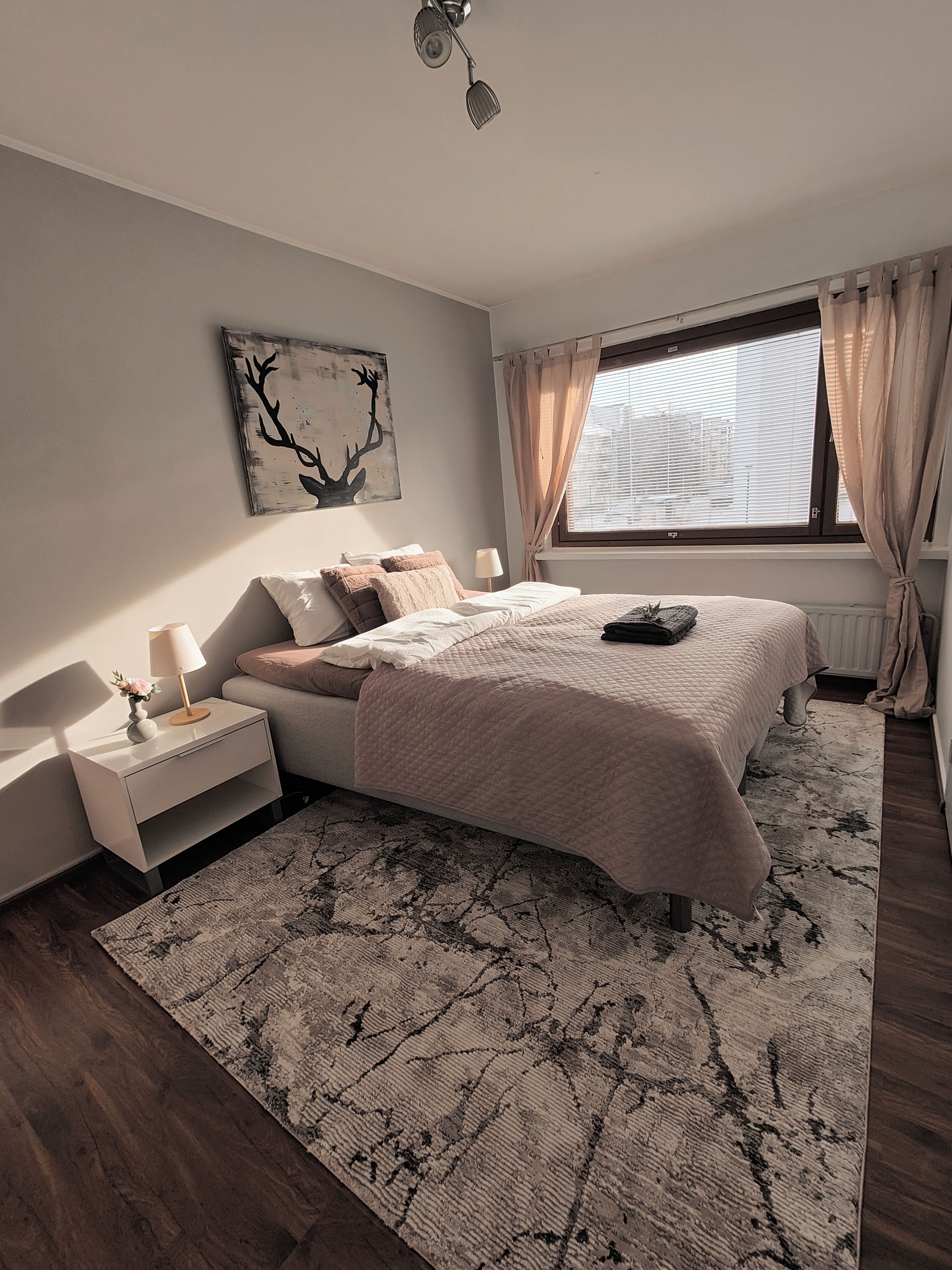 Un dormitorio con cama, mesita de noche, lámpara, obra de arte y vistas al exterior por una ventana.