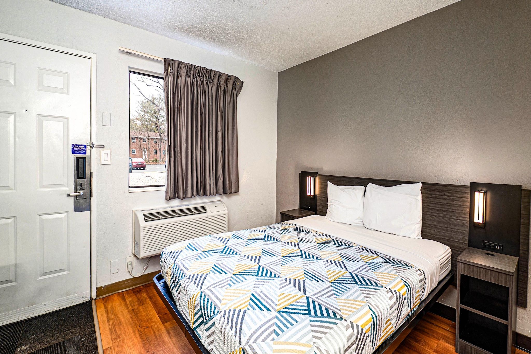 A hotel room with a bed, a window with curtains, an air conditioning unit, and a door with a security lock.