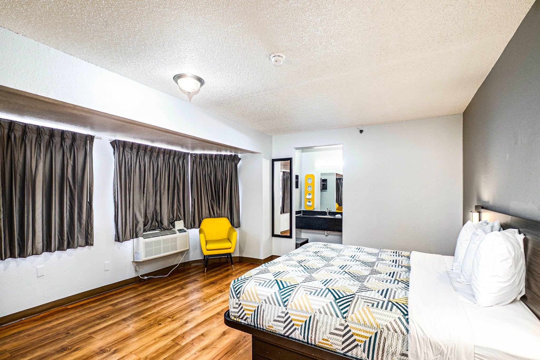 A hotel room with a bed, a chair, a mirror, and a window with curtains.