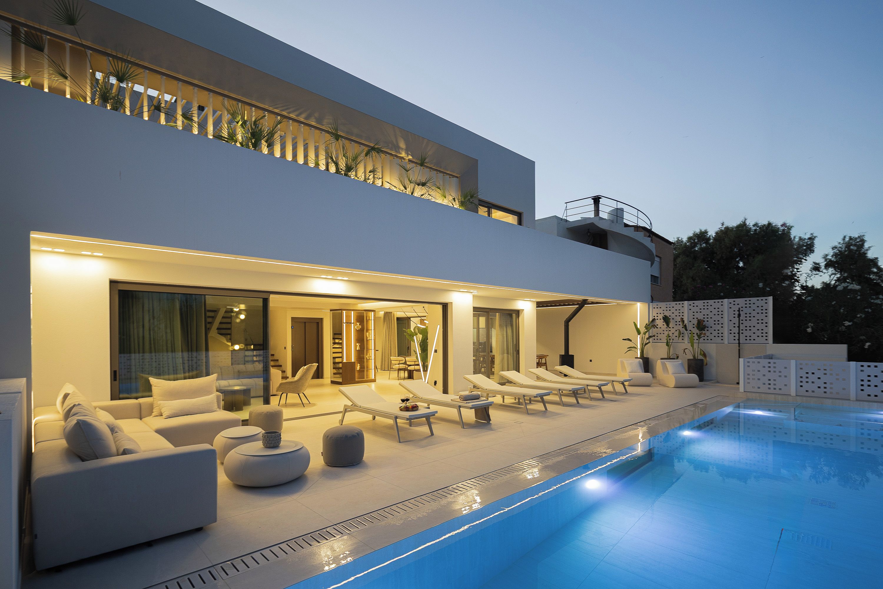 A modern two-story building with a swimming pool, lounge chairs, and a well-lit interior.