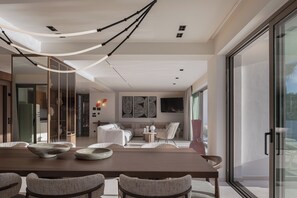 Ein modernes Interieur mit Essbereich, der einen Holztisch und gewebte Stühle umfasst, einem Wohnbereich mit Sofa und Kunstwerken sowie einer Glastür, die nach draußen führt.