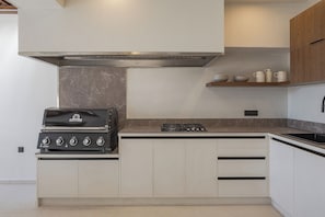Cozinha compacta com fogão a gás embutido, churrasqueira Broil King e prateleira de madeira sobre o balcão.