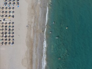 Vista aérea de uma praia com fileiras de guarda-sóis e cadeiras de praia, a costa arenosa e o oceano.