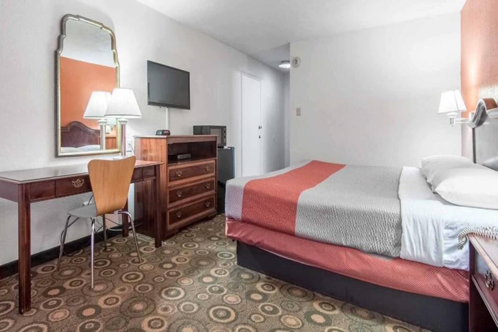 A hotel room with a bed, a desk, a chair, a dresser, a television, and a door.
